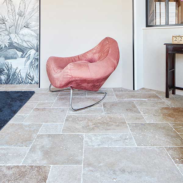 Natural stone flooring in warm beige tones with an opus pattern, featuring a modern pink chair with a chrome base and a black-and-white botanical wallpaper in a stylish interior setting.