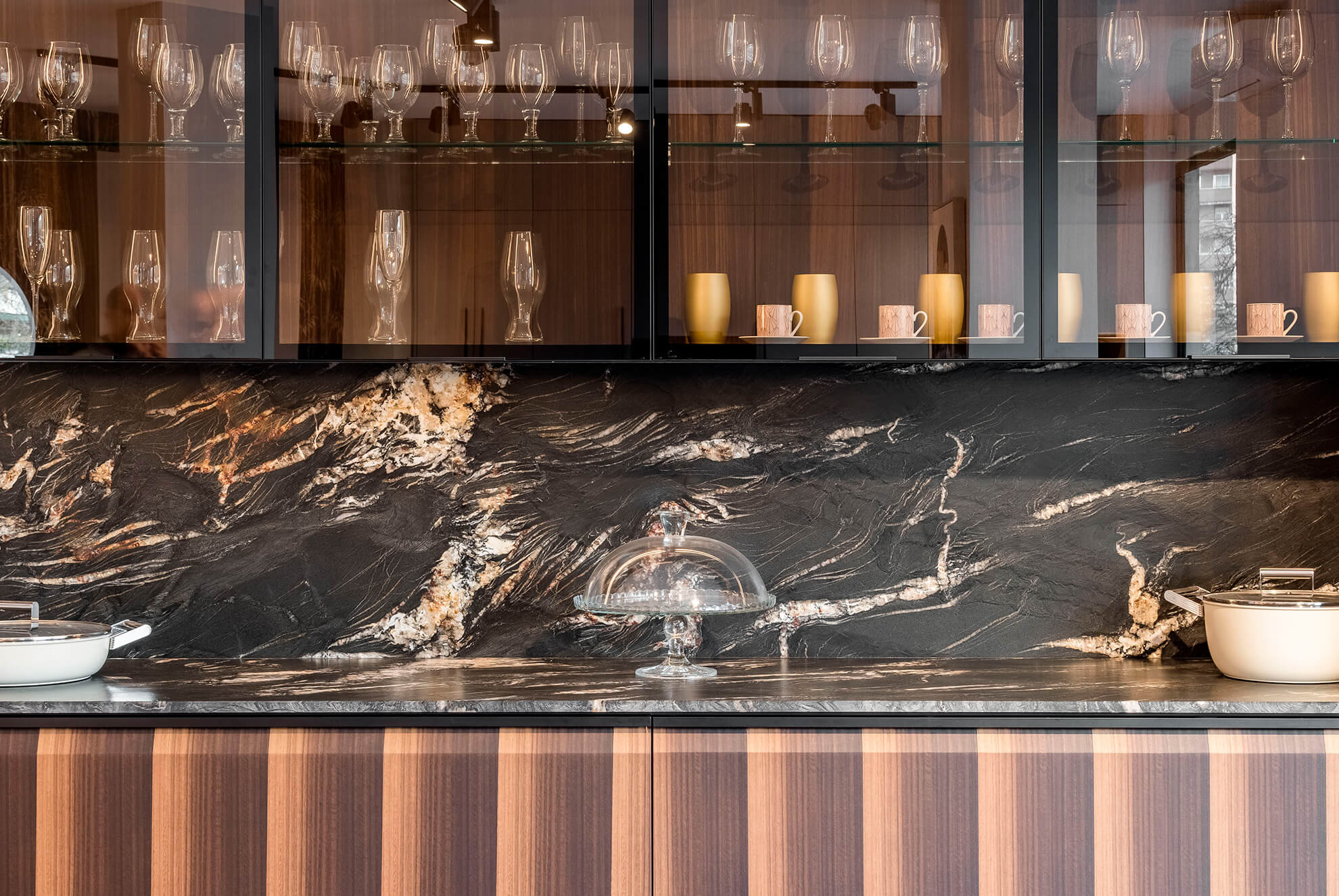 Modern kitchen with a striking black and gold natural stone backsplash, wood cabinetry, and glass display cabinets with integrated lighting.